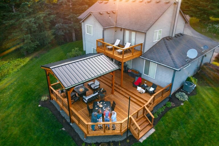 Aerial view of a modern house with a large, multi-level wooden deck. Four people are gathered around a table with food, and lounge chairs are arranged on the deck. The home is surrounded by green lawn and trees.