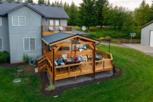 Four people sit around a table on a covered wooden deck attached to a gray house, with a basketball hoop and trees in the background.