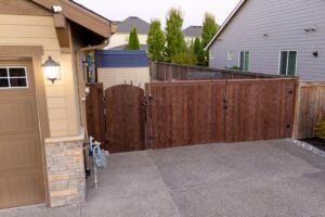 A wooden driveway gate installed between a garage and a house, with a small shed visible in the backyard.