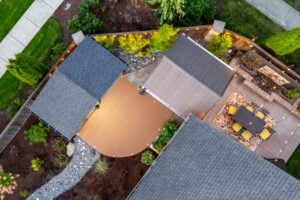 Aerial view of a backyard featuring a curved wooden deck between two building roofs, with an outdoor dining table on a patterned rug nearby, all illuminated by string lights.