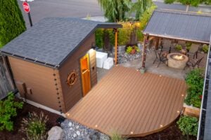 An elevated view of a backyard featuring a wooden sauna with a ships wheel, a curved composite deck, and a stone fire pit area with seating under a pergola.