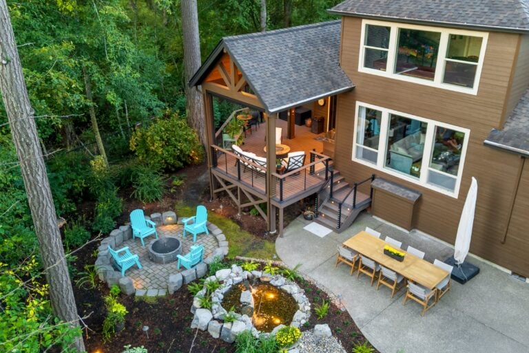 A backyard features a raised wooden deck with seating, a dining area on a concrete patio, a circular fire pit with blue chairs, and a small landscaped pond, surrounded by trees and greenery.