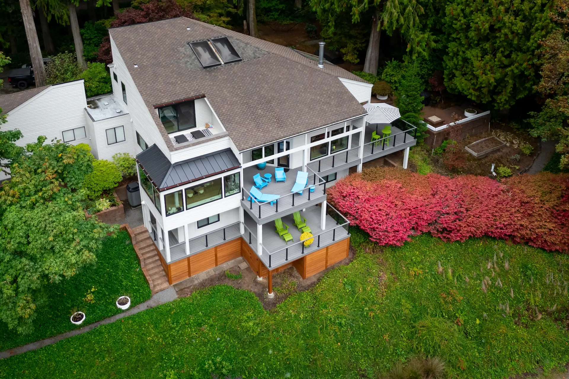 A modern two-story house with multiple balconies, large windows, outdoor seating, and surrounded by lush greenery and red shrubs, viewed from above.