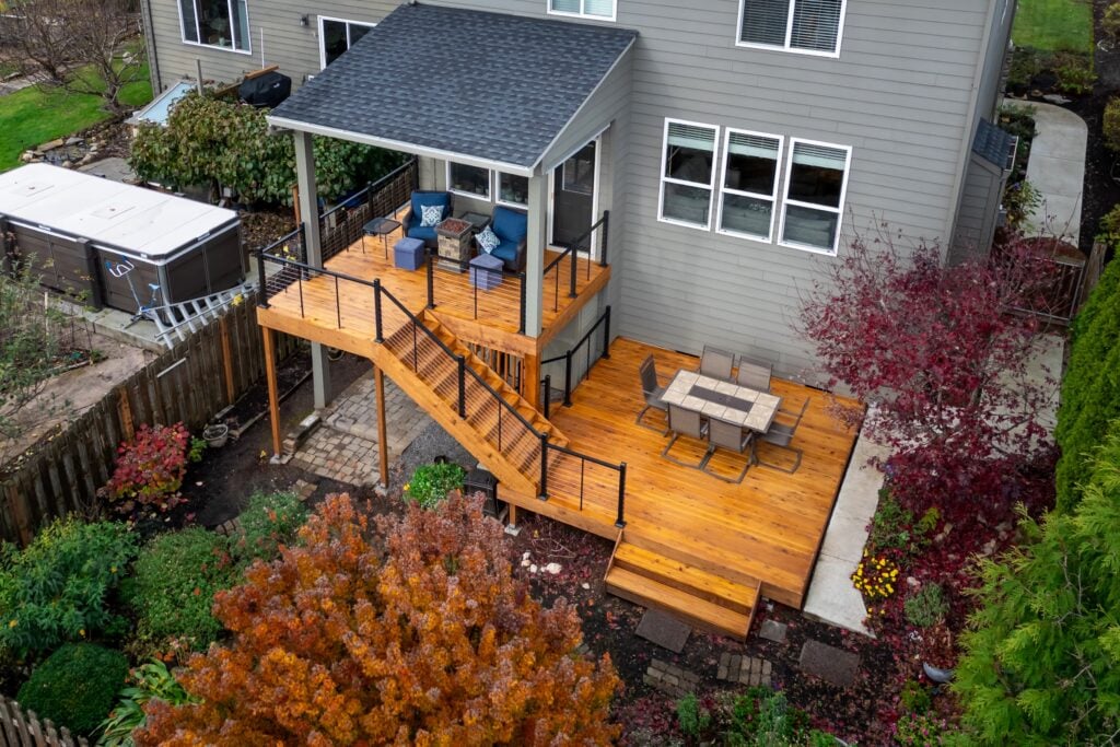 Aerial view of a two-level wooden deck attached to a gray house, with outdoor furniture, stairs, and surrounded by trees and garden beds in autumn.