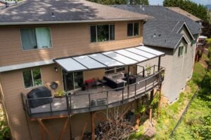 A two-story tan house with a large, elevated deck featuring a covered patio area, outdoor lounge furniture, and a grill.