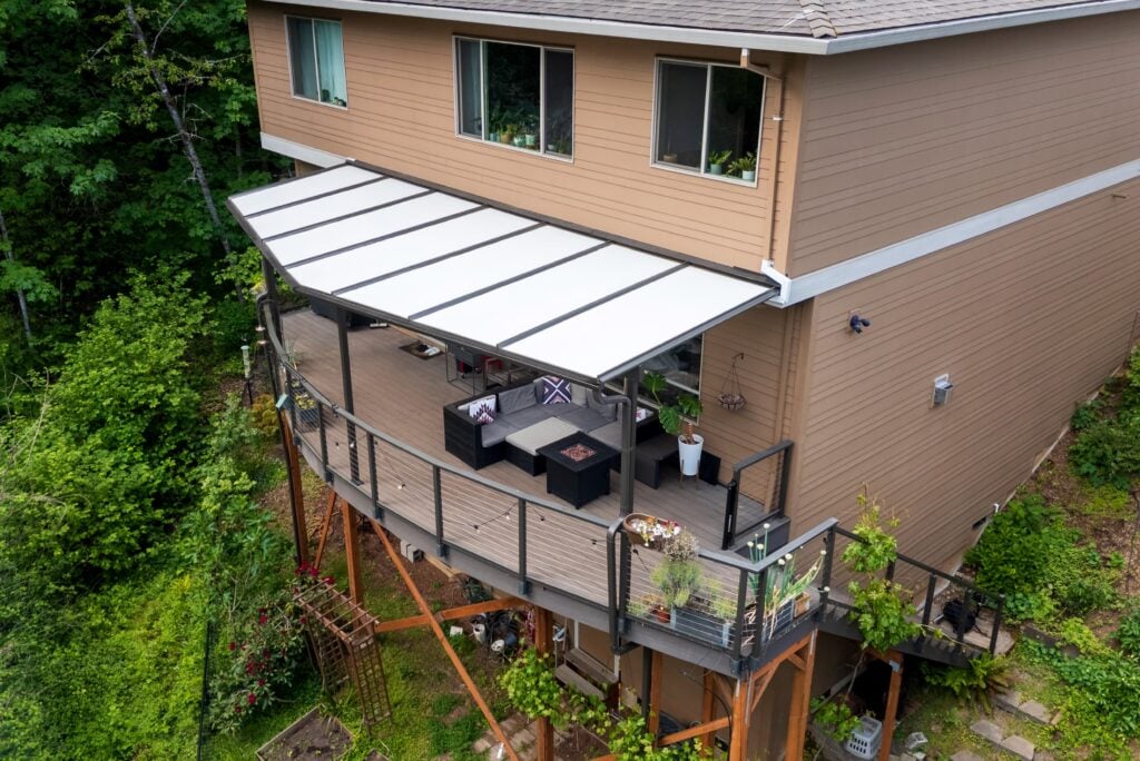Aerial view of a two-story tan house with a large wooden deck featuring a covered seating area and a fire pit, overlooking a wooded hillside.