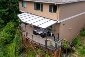 Aerial view of a two-story tan house with a large wooden deck featuring a covered seating area and a fire pit, overlooking a wooded hillside.