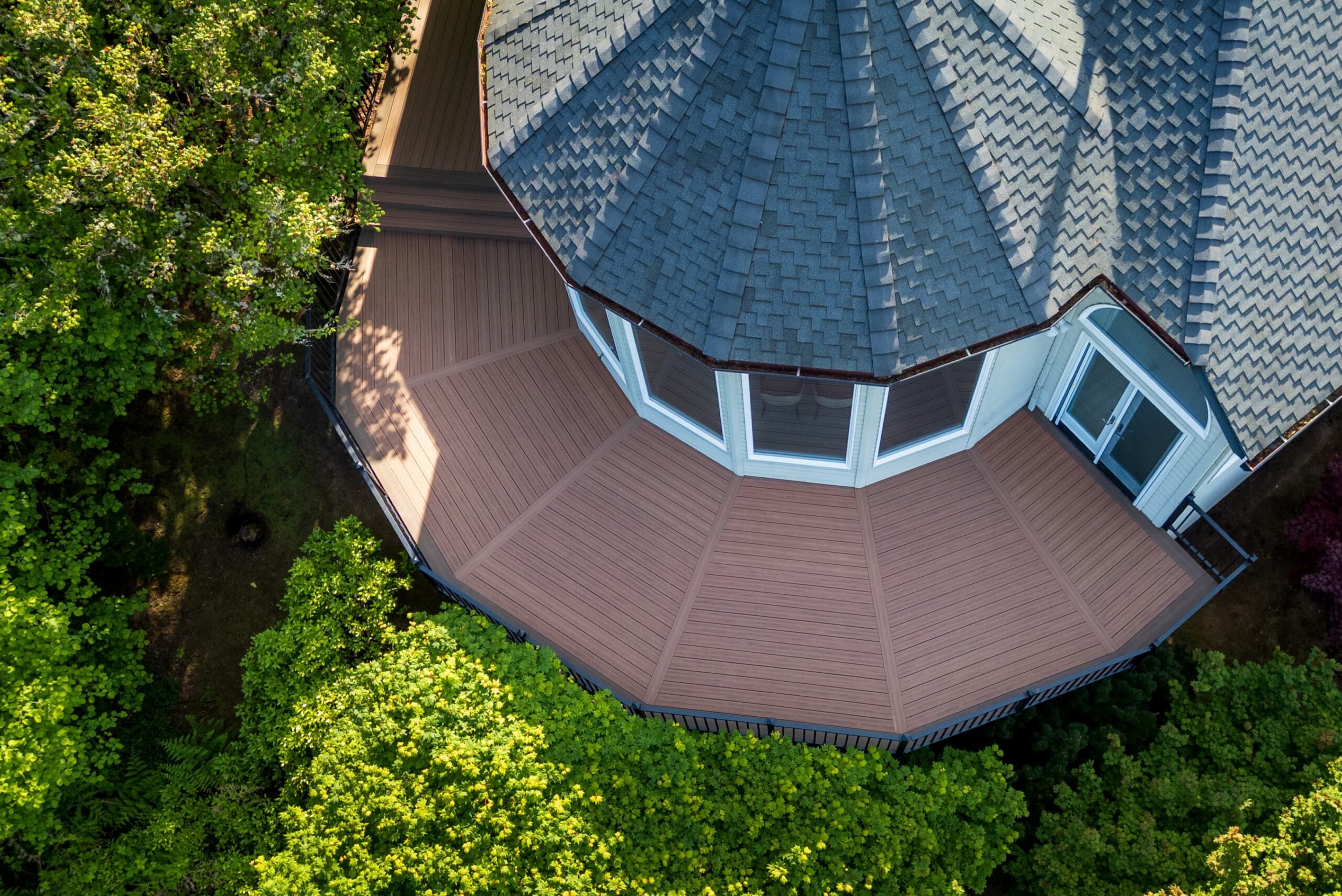 Aerial view of a house with a multi-sided, shingled roof and a spacious, brown deck surrounded by lush green trees. The deck wraps around much of the house, creating a geometric pattern.