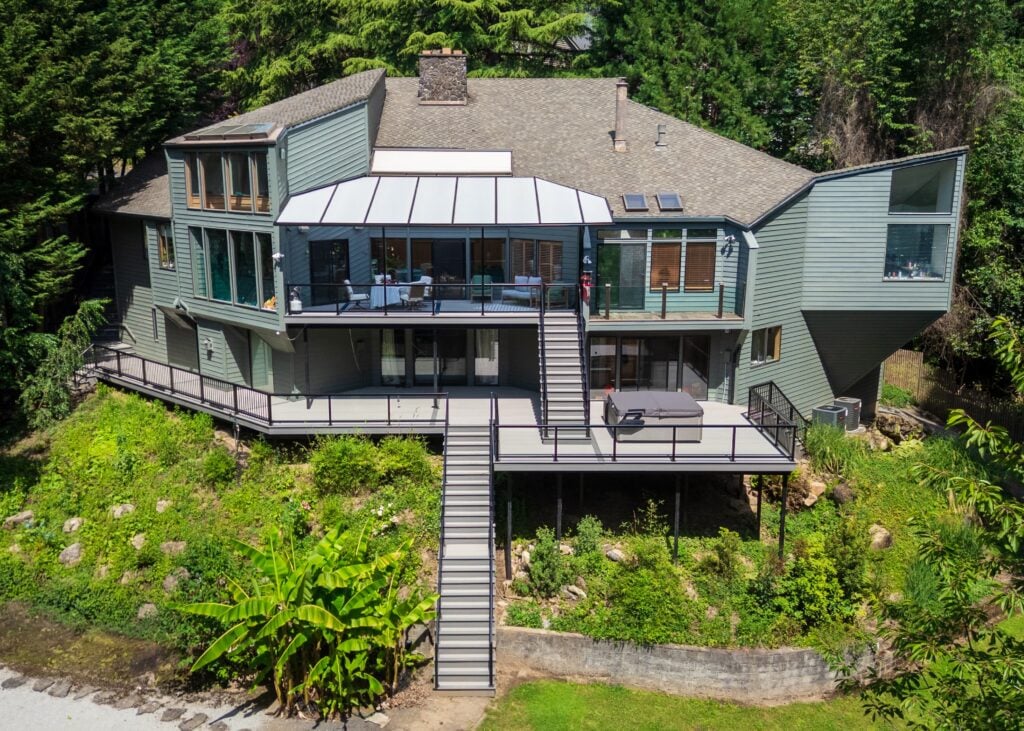 Aerial view of a multi-level green house nestled in a forest, featuring a large deck with a translucent cover, a staircase leading to a lower patio, and a hot tub.