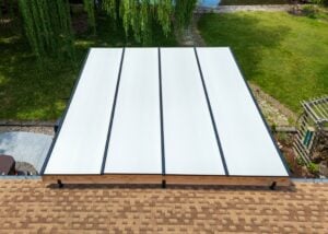 High-angle view of a rectangular patio cover with white panels and black frames attached to a shingled roof, overlooking a backyard lawn.