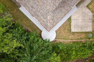 Aerial view of a house corner with a wrap-around deck featuring a semi-circular section, adjacent to a tree-lined yard and concrete patio.