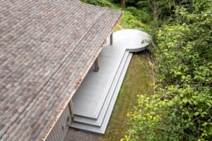 High-angle view of a grey multi-level deck with a circular platform extending from the side of a house into a wooded yard.
