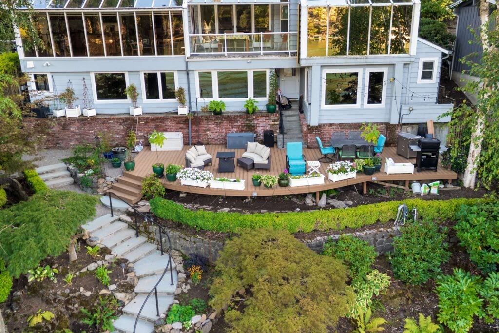 Aerial view of a tiered backyard featuring a wooden deck furnished with patio seating and dining sets, situated behind a multi-story house with a brick base and glass-enclosed upper levels.