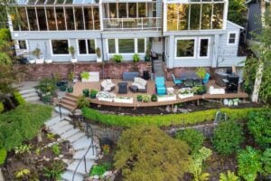 Aerial view of a tiered backyard featuring a wooden deck furnished with patio seating and dining sets, situated behind a multi-story house with a brick base and glass-enclosed upper levels.