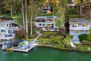 An elevated view of three lakefront houses nestled in a wooded hillside, featuring a shared dock with lounge chairs and a covered boat slip.