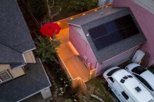 Aerial view of a backyard wooden deck illuminated by ground lighting next to a pink house with solar panels on the roof.