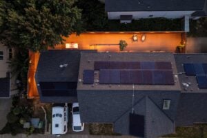 Aerial view of a house at twilight, featuring solar panels on the roof and a well-lit wooden deck in the backyard with lounge chairs and a dining table.