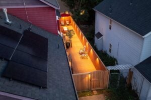 An elevated night view of a narrow residential deck illuminated by wall lights, featuring patio furniture and a wooden gate.
