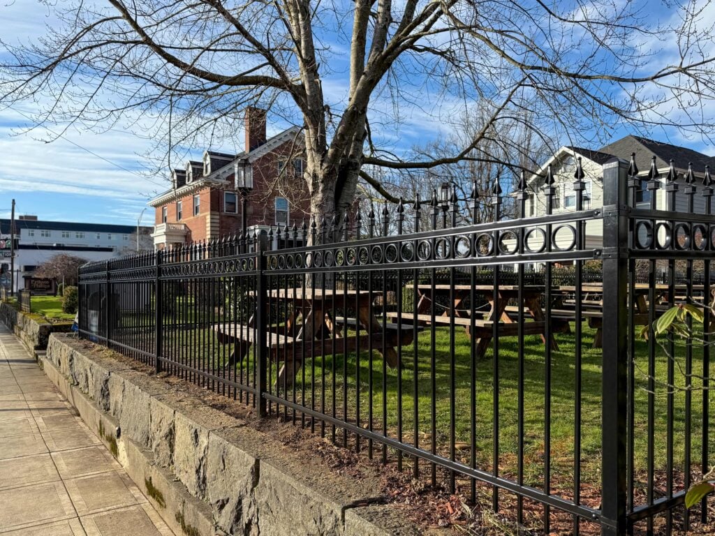 A black metal fence surrounds picnic tables and a large tree on green grass, with a red brick building and a blue sky in the background.