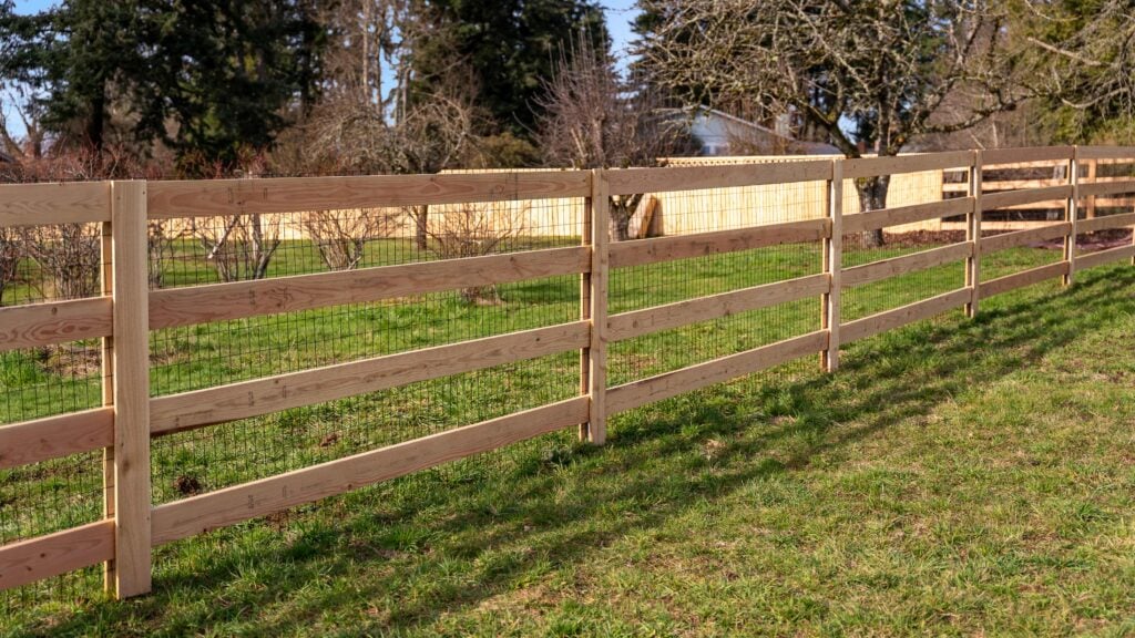A wooden split-rail fence with wire mesh runs through a grassy yard, with trees, shrubs, and another fence in the background under a clear sky.
