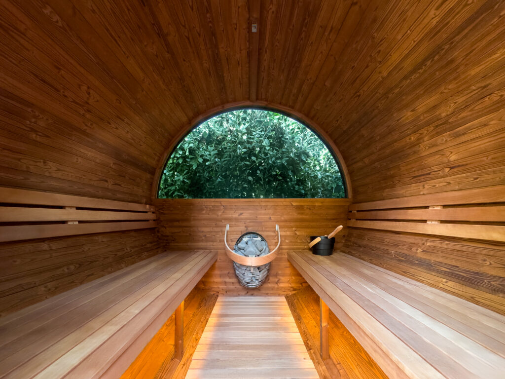 Interior of a barrel-shaped wooden sauna with two benches, a sauna heater, and a bucket with a ladle. A large window at the back reveals greenery outside.