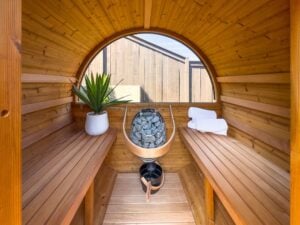 Inside of a wooden barrel sauna with two bench seats, a heater filled with stones, a potted plant, and folded white towels.