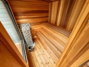 Interior of a cedar sauna featuring tiered wooden benches and a vertical heater filled with stones next to a water bucket.