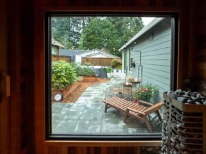 View through a window of a patio with stone pavers, a wooden lounge chair, planter boxes, and a sauna heater in the foreground.