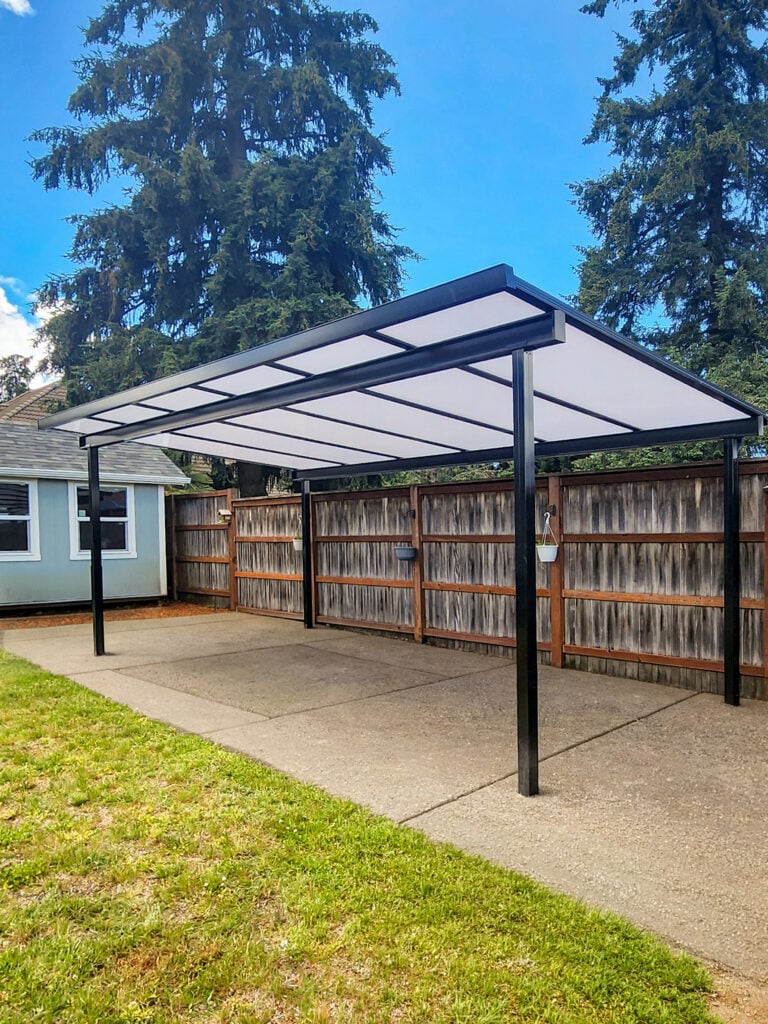 A modern metal carport with a translucent roof stands on a concrete driveway next to a wooden fence and a small house, with grass and tall trees in the background under a blue sky.