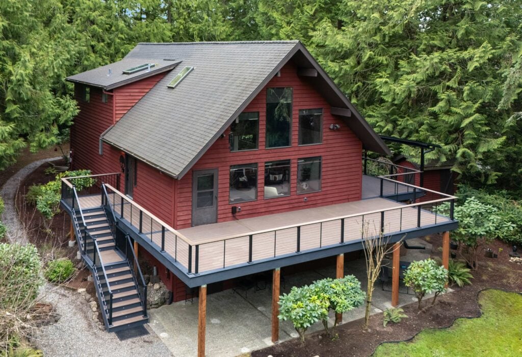 A two-story red cabin with large windows and an upper-level wraparound deck, surrounded by dense green trees and shrubbery.