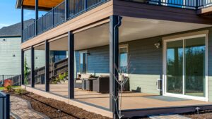 A covered ground-level deck with brown composite flooring, black support posts, and patio furniture, located beneath a raised second-story deck.