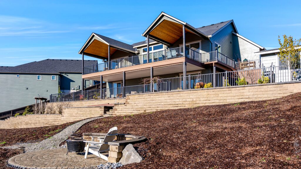 A two-story house with two covered balconies overlooking a backyard with tiered retaining walls and a circular stone patio featuring a white Adirondack chair and a fire pit.