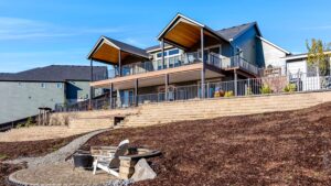 A two-story house with two covered balconies overlooking a backyard with tiered retaining walls and a circular stone patio featuring a white Adirondack chair and a fire pit.