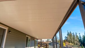 A covered patio area attached to a house, featuring a light-colored tongue-and-groove ceiling and a view of trees and a blue sky.