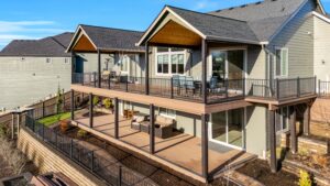 A two-story house with a large upper deck featuring outdoor dining furniture and a lower covered patio with lounge seating, both enclosed by black metal railings.