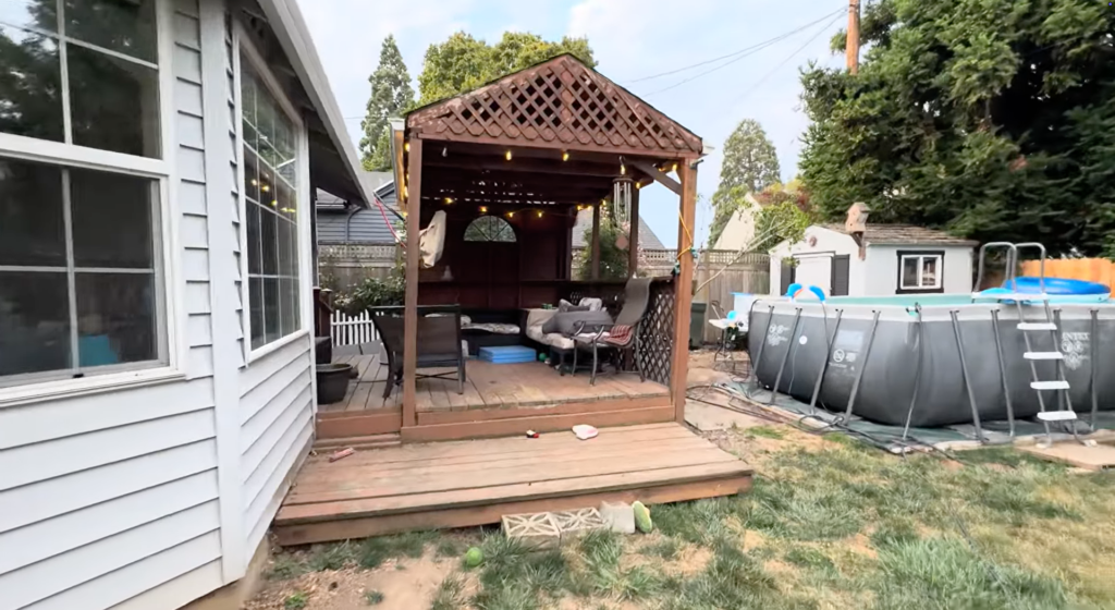 A backyard with a wooden gazebo containing outdoor furniture, string lights, and toys. To the right, there is an above-ground pool with a ladder. The scene includes grass, a shed, and tall trees.