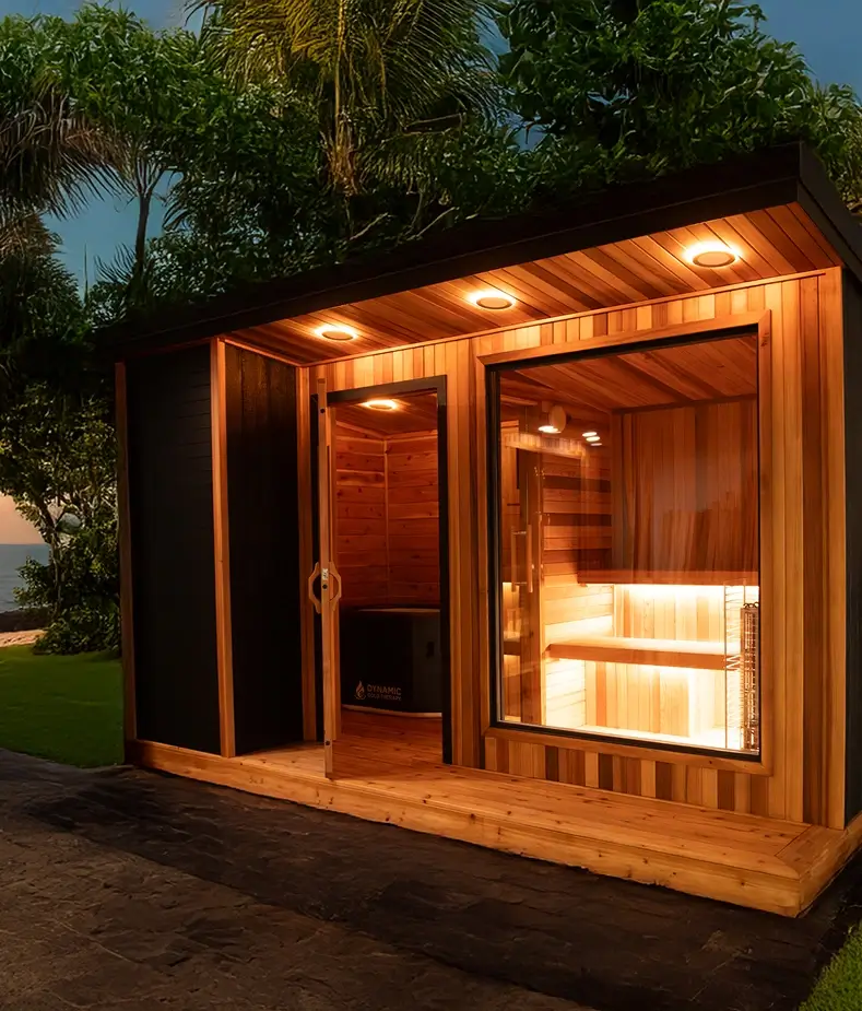 A modern outdoor sauna with warm interior lighting, wood-paneled walls, large windows, and a glass door, set in a lush, tropical garden during the evening.