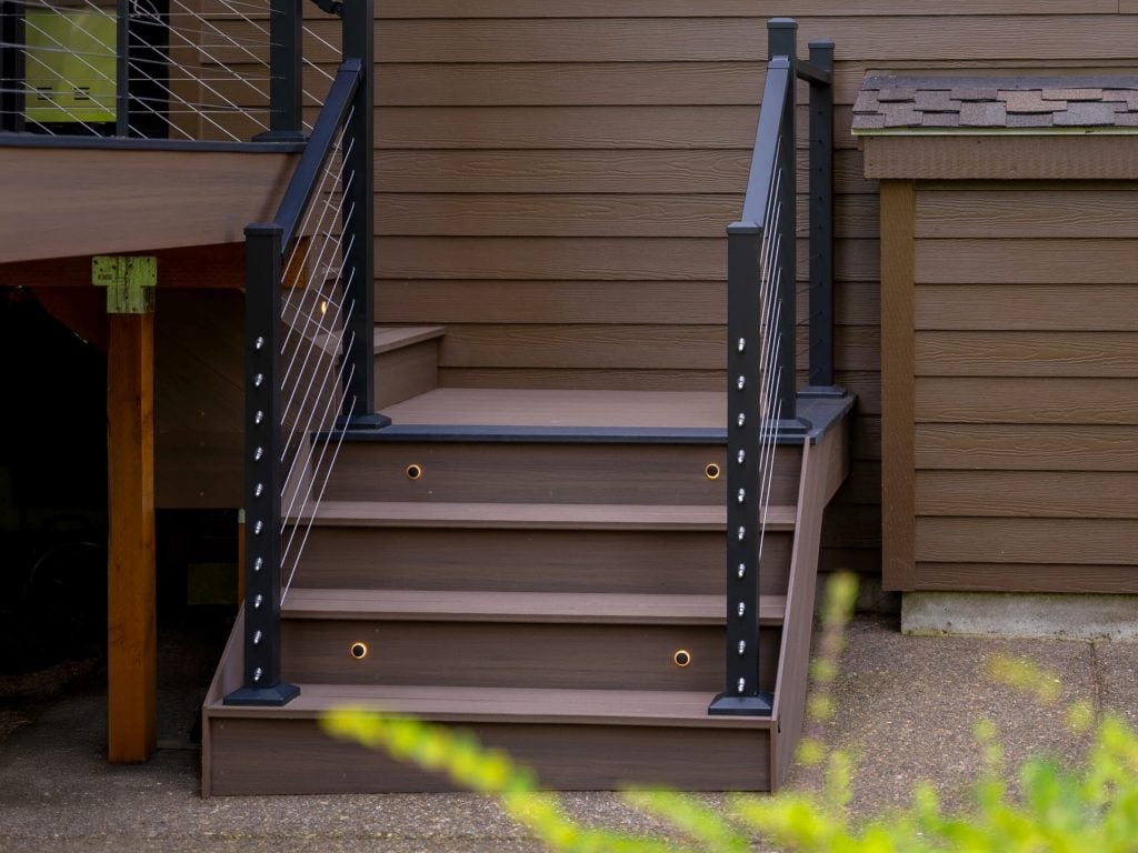 A small set of outdoor stairs with brown wooden steps and black metal railings with horizontal cables, attached to the side of a brown house. Shrubs are partially visible in the foreground.