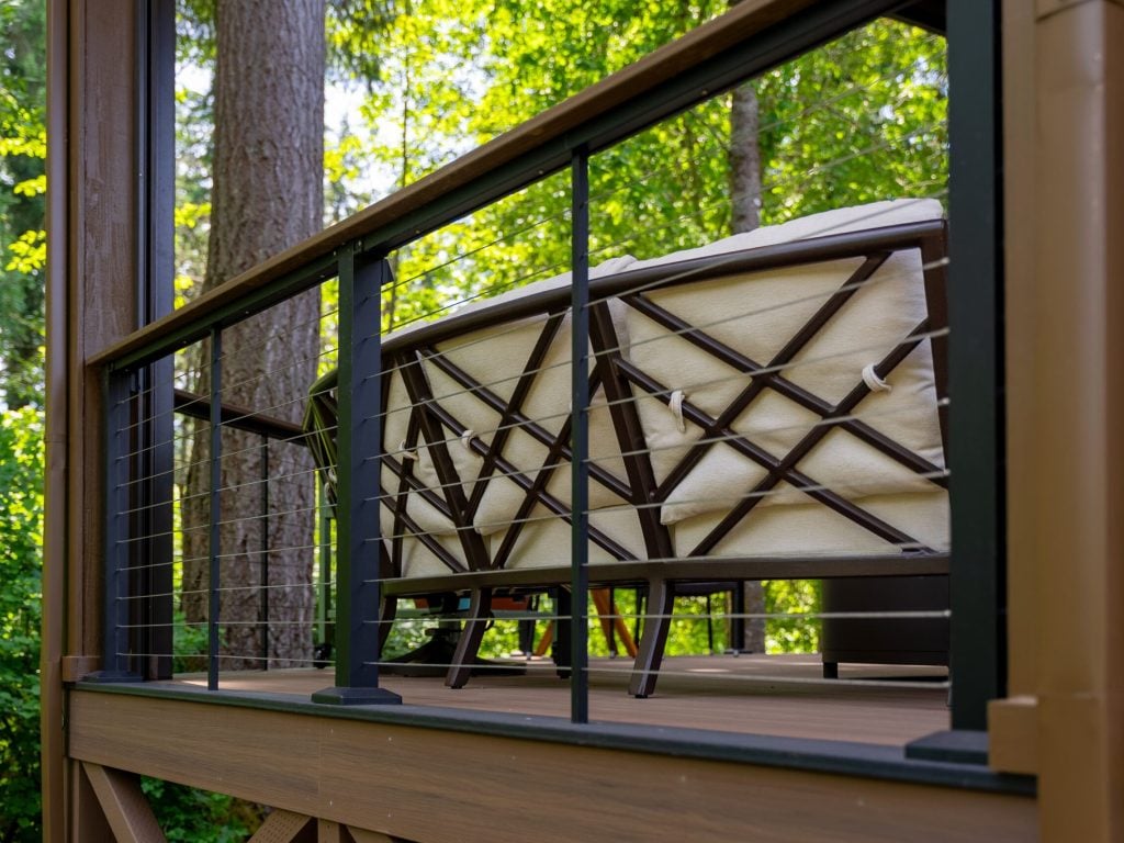 Close-up of a modern deck railing with metal cables and brown posts, framing a cushioned outdoor chair. Lush green trees and foliage are visible in the sunny background.