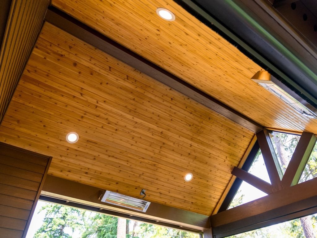 Angled view of a modern wooden ceiling with recessed lights and a triangular beam structure, seen from below, with large windows revealing green trees outside.