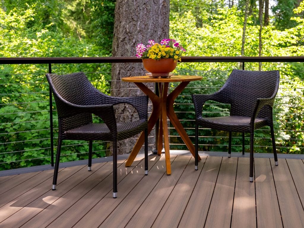 Two black wicker chairs and a small wooden round table with a potted pink flower, set on a wooden deck surrounded by lush green trees and foliage.