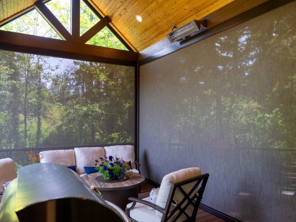 A screened-in patio with cushioned chairs, a round table with a flower arrangement, a grill in the foreground, and trees visible through the mesh screens.