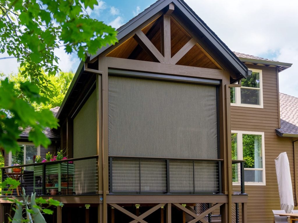 A modern two-story house with brown siding features a covered deck with a retractable screen, metal railing, and large windows. Green trees and a patio umbrella are visible nearby under a partly cloudy sky.