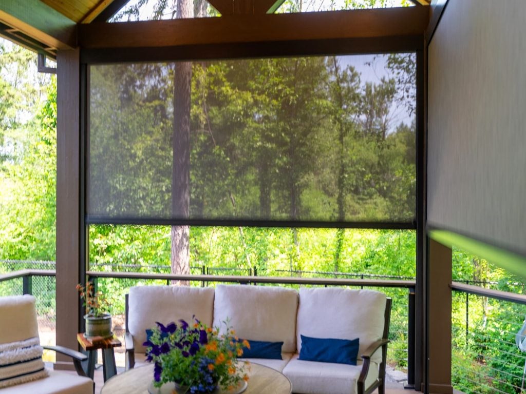 A screened-in porch with a wood ceiling, modern outdoor furniture with cream cushions, a round coffee table with a flower arrangement, and a view of green trees outside.