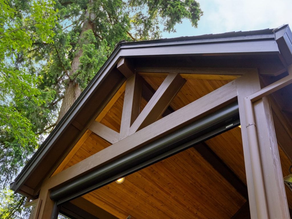 Close-up view of the wooden roof structure of an outdoor building or pavilion, with exposed beams and natural wood ceiling, surrounded by tall green trees.