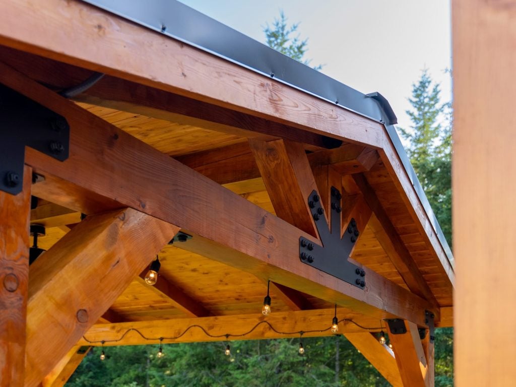 Close-up of a wooden pergola roof with metal brackets and string lights hanging underneath, set outdoors with green trees visible in the background.