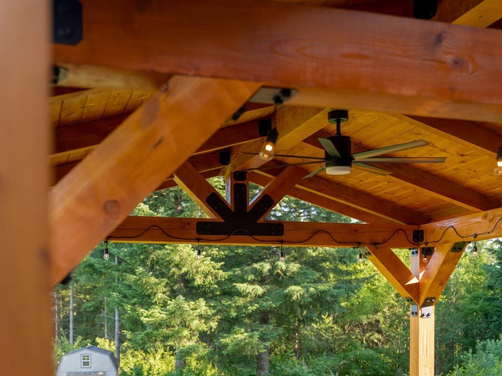 A wooden outdoor pavilion with exposed beams, hanging lights, and a ceiling fan, set against a backdrop of green trees and a small shed in the distance.