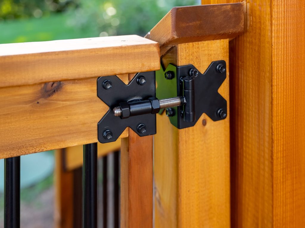 Close-up of a black metal hinge attached to two sections of a wooden fence or railing, connecting the corner where two polished wooden posts meet. The background is blurred greenery.