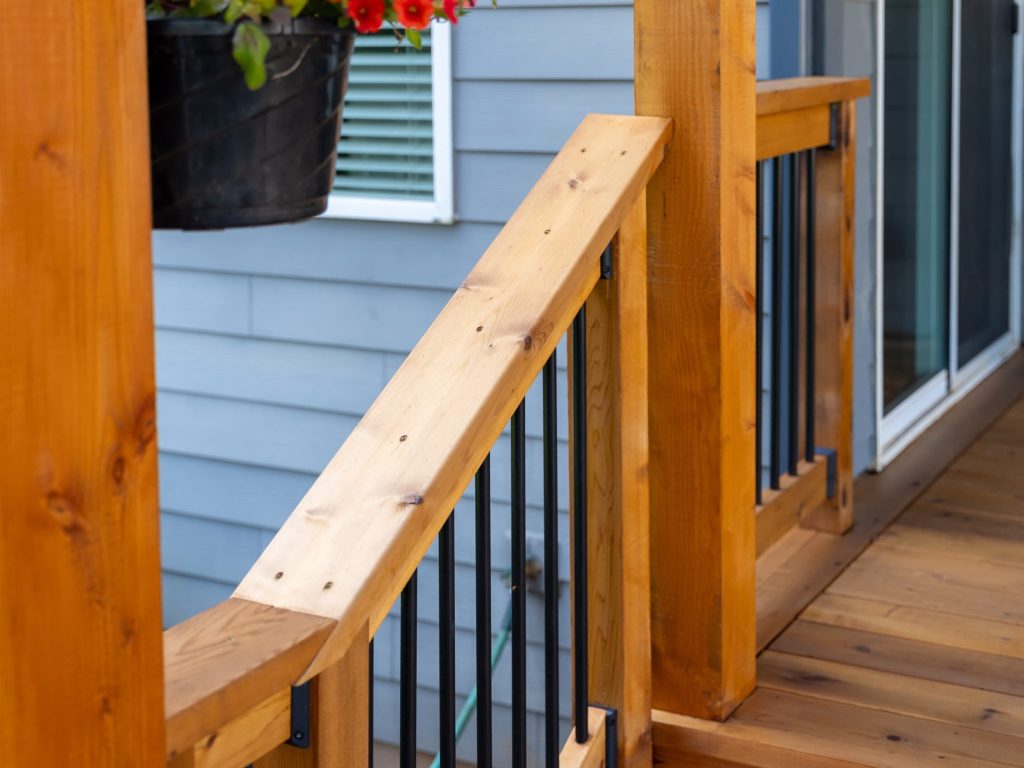 Close-up of a wooden porch railing with vertical metal balusters, next to light blue house siding and a sliding glass door. A hanging planter with red flowers is visible in the upper left corner.