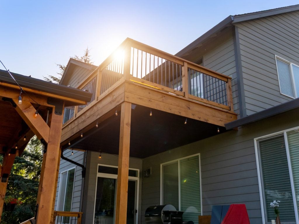 A two-story gray house with a wooden balcony and string lights, basking in sunlight. There’s a covered patio below, large windows, and a grill on the deck. Trees and a clear blue sky are in the background.